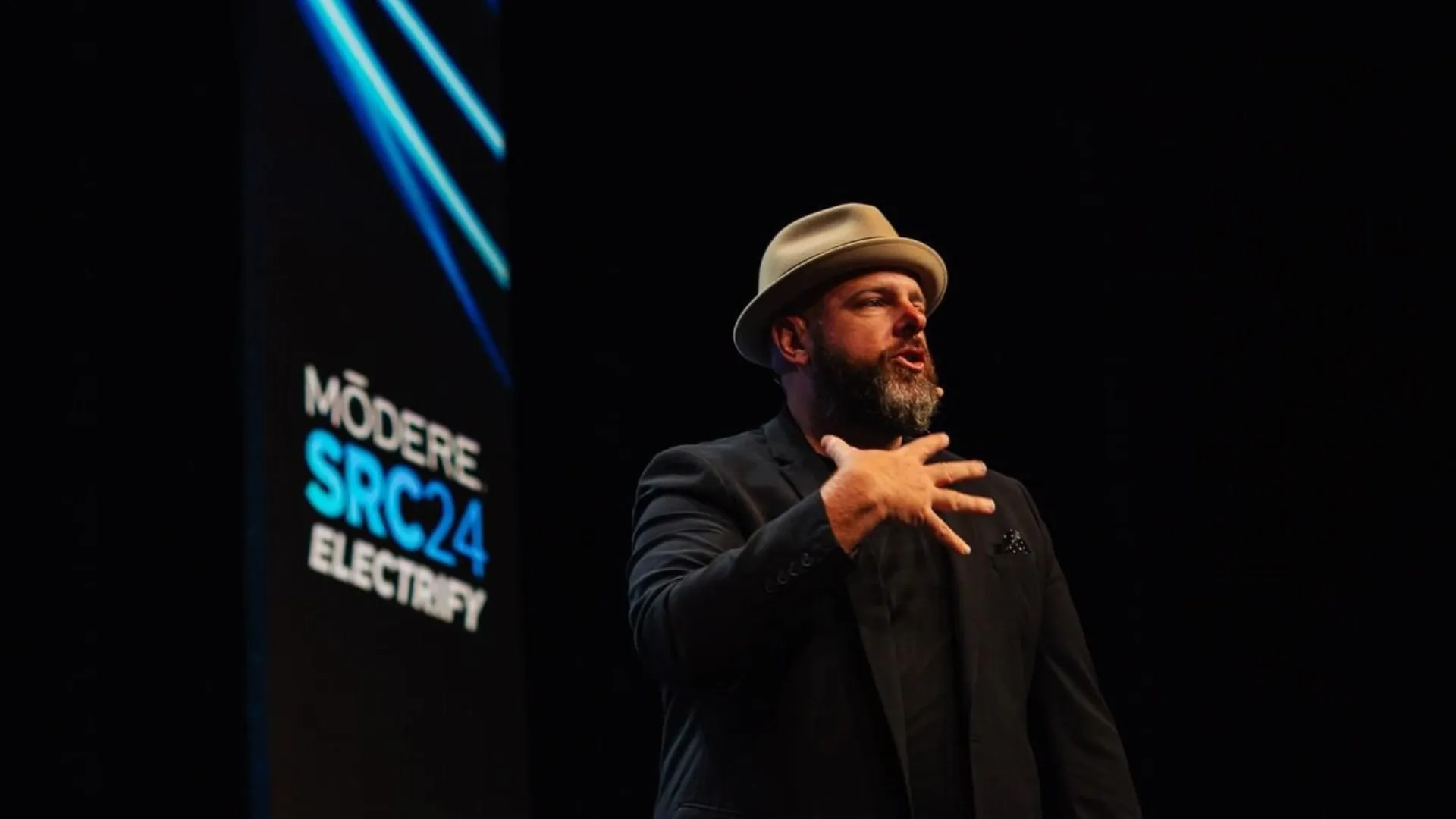 Brett on a dark stage in a fedora and black jacket, mid-sentence, hand on his chest